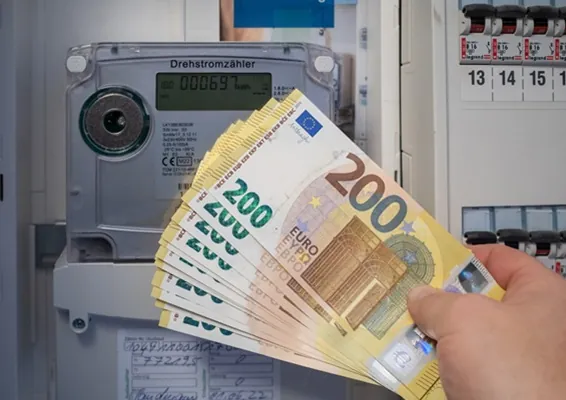 Una mano tiene una mazzetta di banconote davanti ad un contatore della luce